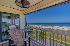 A balcony with a view of the beach, a fan, and a table with chairs.