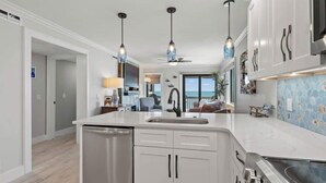 A modern kitchen with white cabinetry, a stainless steel dishwasher, and a large island with a sink. There are pendant lights above the island and a ceiling fan in the background. - Ocean Forest Villas C310 - Oceanfront (Myrtle Beach)