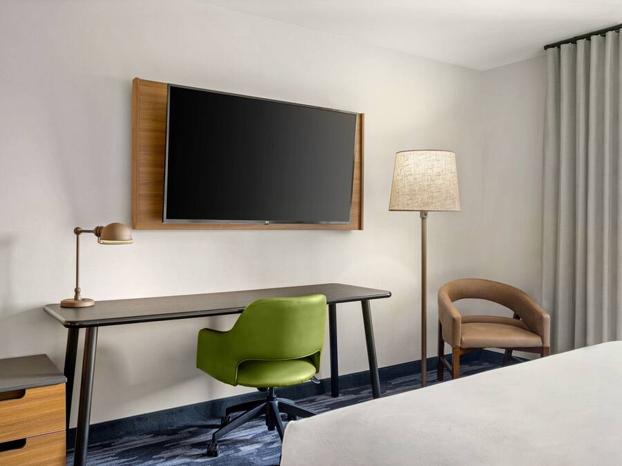 A hotel room with a bed, a desk, a green chair, a brown chair, a television, and a lamp.