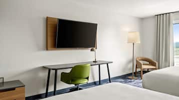 Een hotelkamer met een bed, een bureau met stoel, een aan de muur bevestigde televisie, een vloerlamp en een raam met gordijnen.