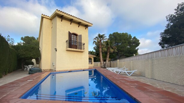 Una piscina con solárium de baldosas, un pequeño edificio con balcón y palmeras al fondo.