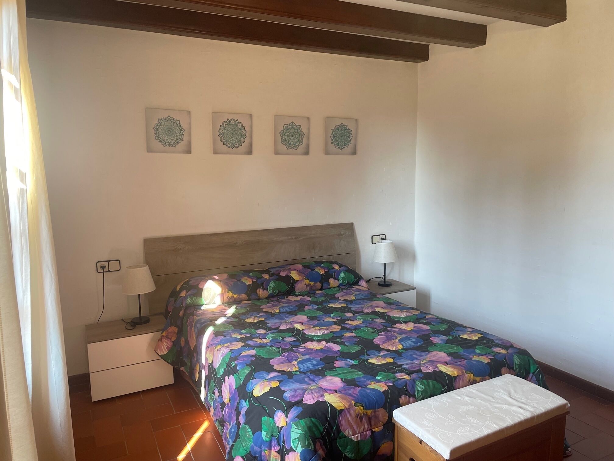 Ein Schlafzimmer mit einer blumigen Tagesdecke, einem hölzernen Kopfende, einem Nachttisch mit Lampe und einer kleinen Bank.
