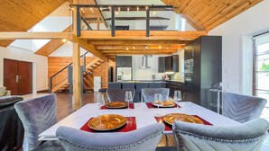 A modern loft-style dining area with a high ceiling, exposed wooden beams, and a kitchen in the background. - Luxury Log Retreat, King Bed, Games Room (Bayfield)