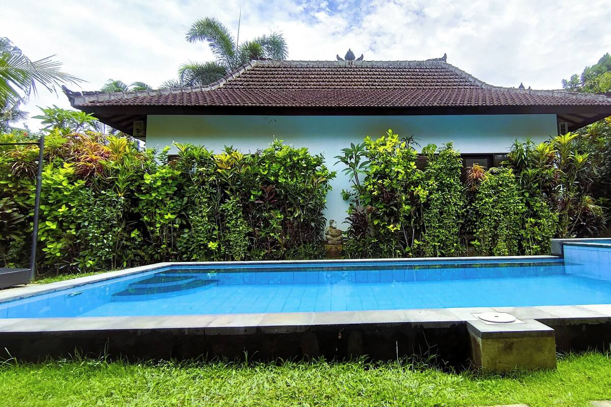 A swimming pool with a tiled deck, surrounded by lush greenery and a traditional building with a tiled roof.