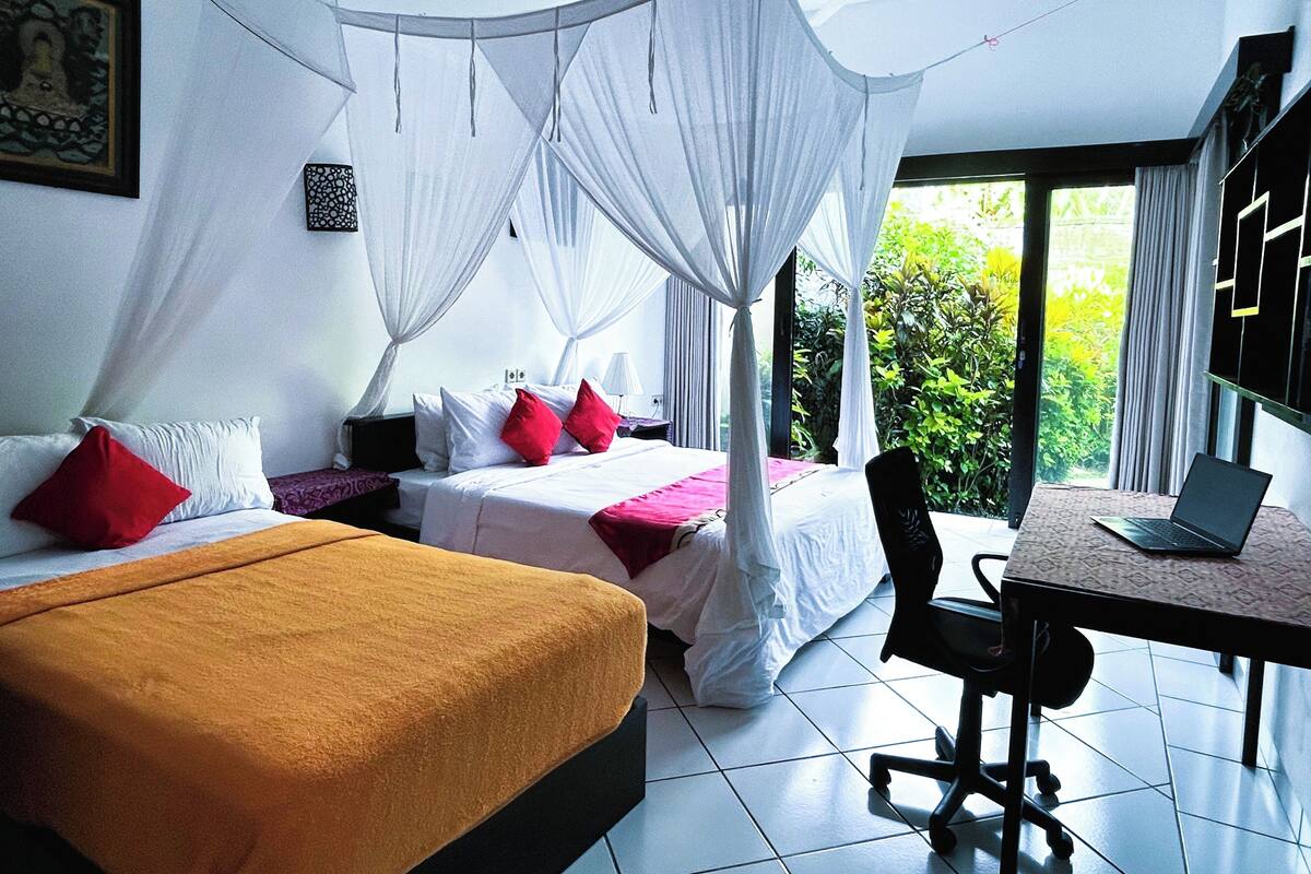 A room with two beds, a desk with a laptop, and a view of greenery outside.