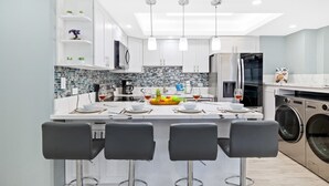 Una cocina moderna con armarios blancos, una isla central con taburetes y una lavadora, además de un comedor con mesa para cuatro.