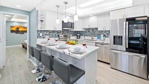 Una cocina moderna con armarios blancos, una isla central con taburetes de bar, electrodomésticos de acero inoxidable e iluminación colgante.