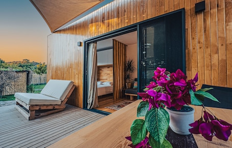 A modern wooden cabin with a wooden deck, a lounge chair, a bedroom with a bed and a window, and a potted plant with purple flowers.
