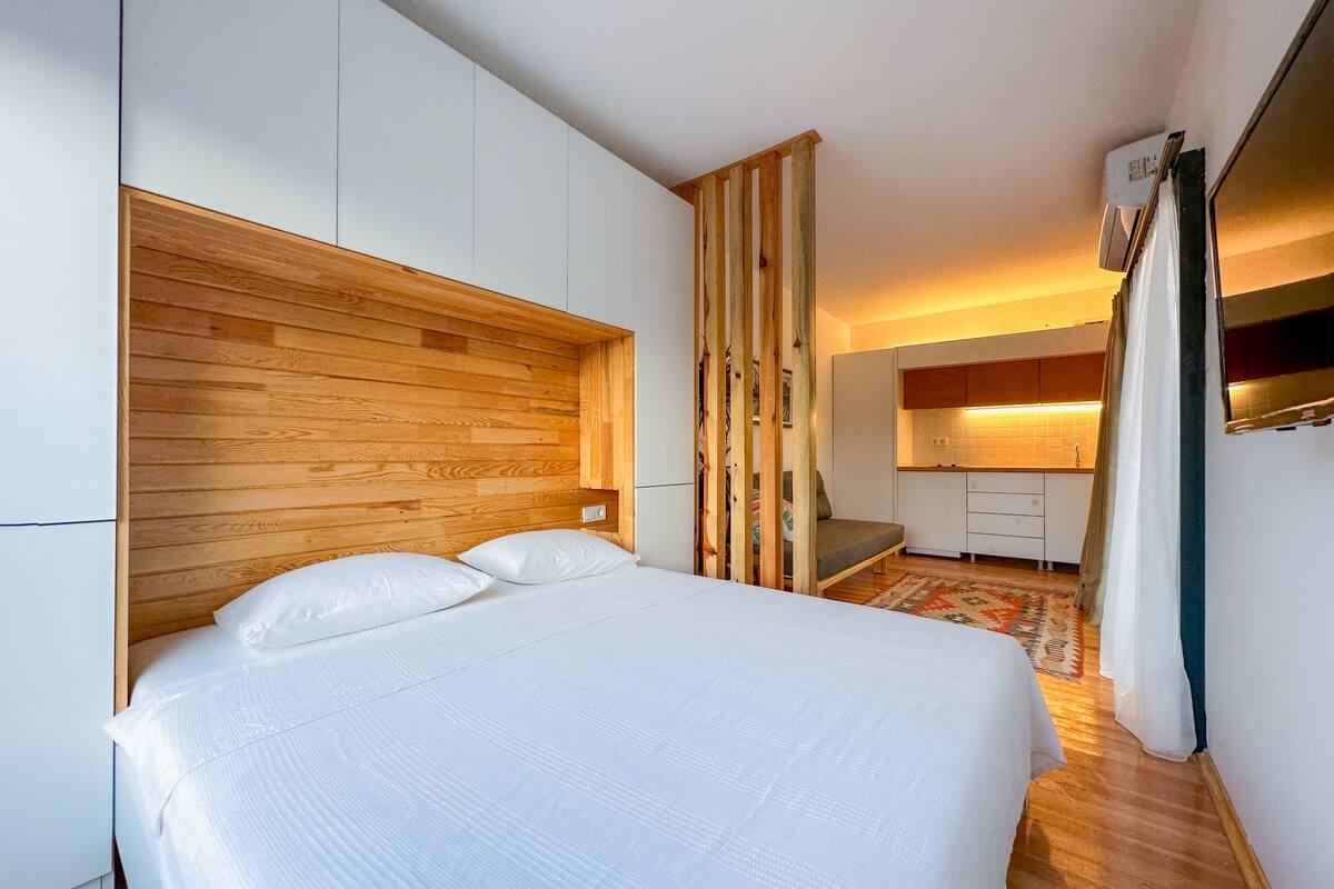 Un dormitorio moderno con cabecera de madera, cama con ropa de cama blanca, una pequeña cocina integrada al fondo y una alfombra estampada en el suelo.