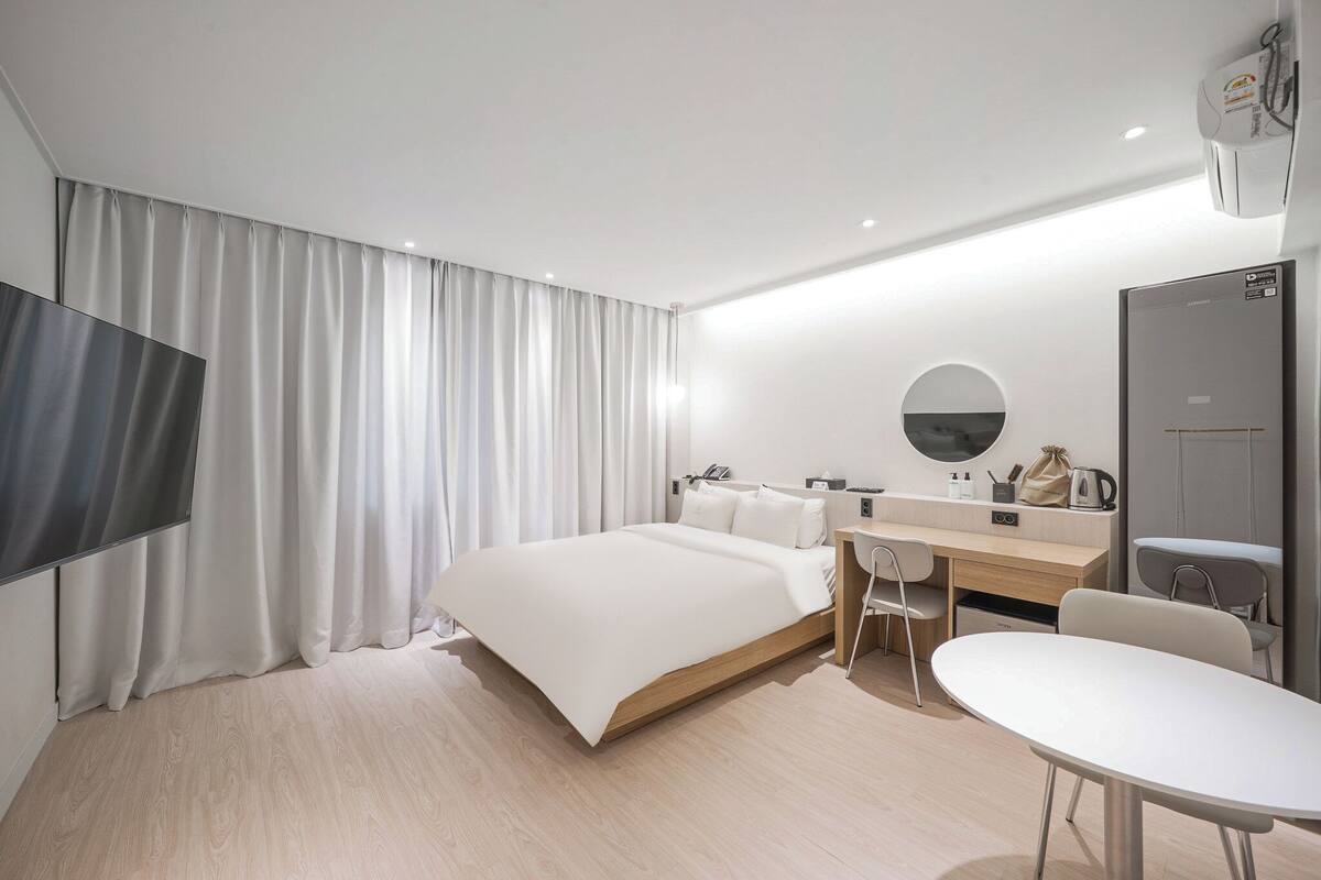 A modern hotel room with a bed, a desk, a chair, a TV, a mirror, and a wardrobe.