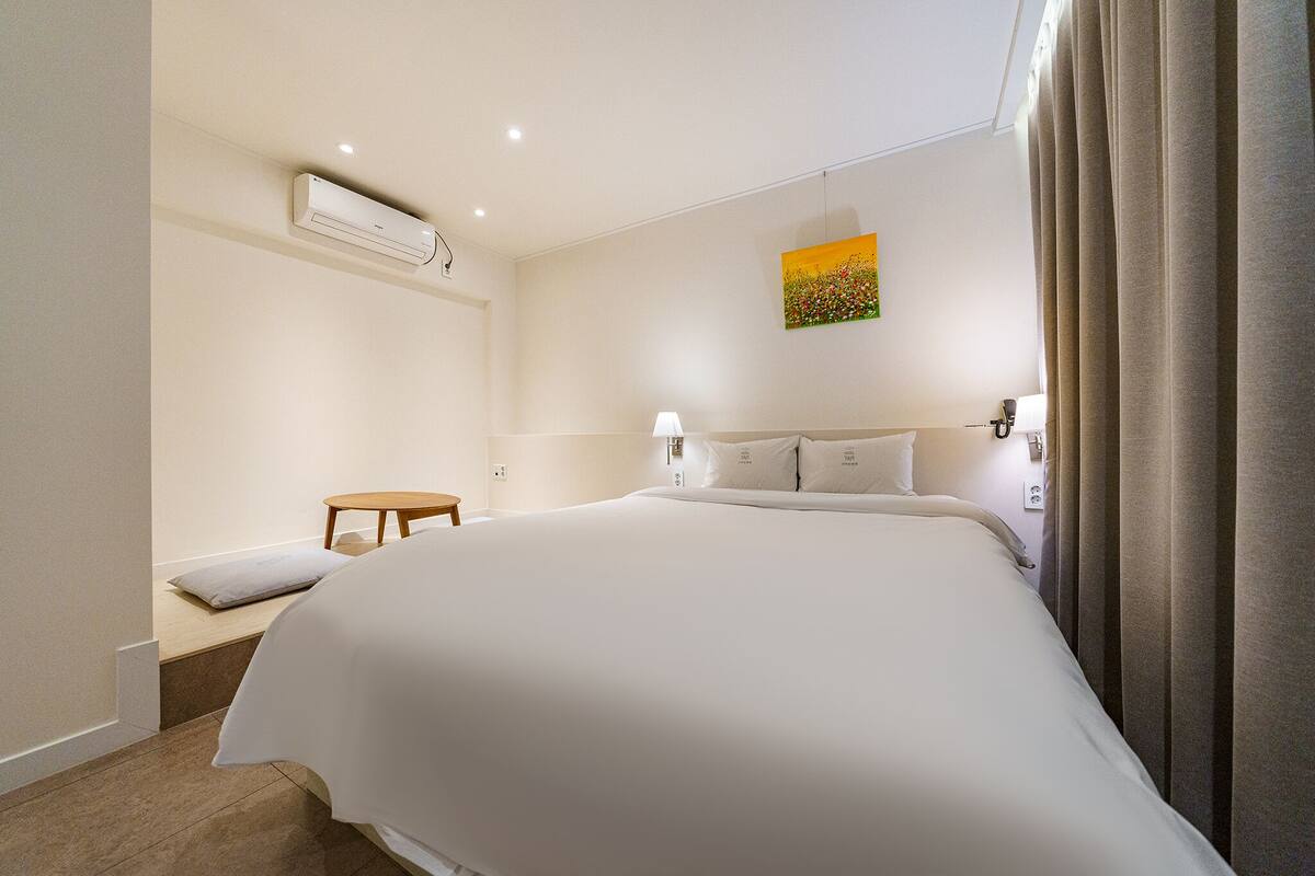 A hotel room with a large bed, bedside lamps, a small table, an air conditioning unit, and a painting on the wall.