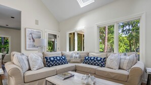 A modern living room with a beige sectional sofa, a coffee table, and a large window with a view of greenery. - Premier Golfside Getaway | Coastal Gallery (Kiawah Island)