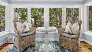 A room with wicker chairs, a round table, and a view of trees through large windows. - Premier Golfside Getaway | Coastal Gallery (Kiawah Island)
