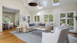 A spacious living room with a large sectional sofa, a coffee table, and a dining area with a table and chairs. There are large windows providing natural light and views of the outdoors. - Premier Golfside Getaway | Coastal Gallery (Kiawah Island)