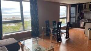 A modern living room with a glass coffee table, a sofa, and a dining area with chairs and a table. Large windows offer a view of the outdoors. - Modern 2 bed apartment with stunning seaside view (Leith)
