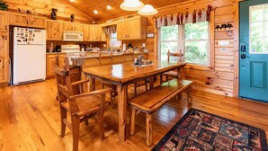 Una cabaña de madera con cocina, que incluye una mesa de madera grande, sillas, un banco y un refrigerador.