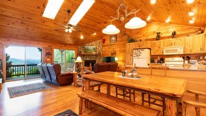 Interior de una cabaña de madera con una gran mesa de comedor, bancos, chimenea y zona de cocina.