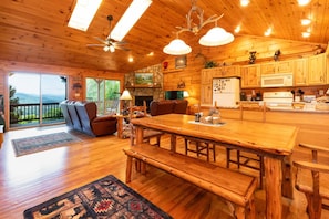 A wooden cabin interior with a large dining table, benches, a fireplace, and a kitchen area.