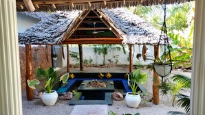A covered outdoor seating area with a thatched roof, surrounded by potted plants and a small water feature.