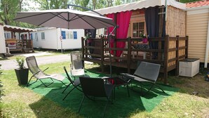 Ein überdachter Außenbereich mit Stühlen und Tisch, umgeben von einer Holzterrasse und einem kleinen Gebäude mit rotem Vorhang.