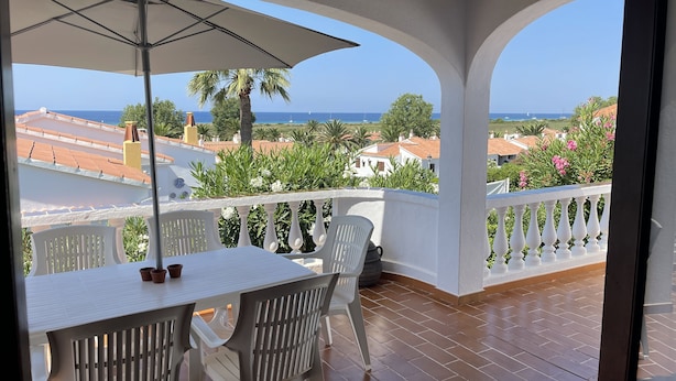 A balcony with a table and chairs, a white umbrella, and a view of the sea and buildings. - 3 Bedroom Villa, San Jaime, Son Bou (Son Bou)