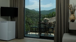 A room with a TV, a small fridge, a dining area with chairs and a table, and a large sliding glass door leading to a balcony with a view of green hills. - Kaktus Suites Hotel (Kas)