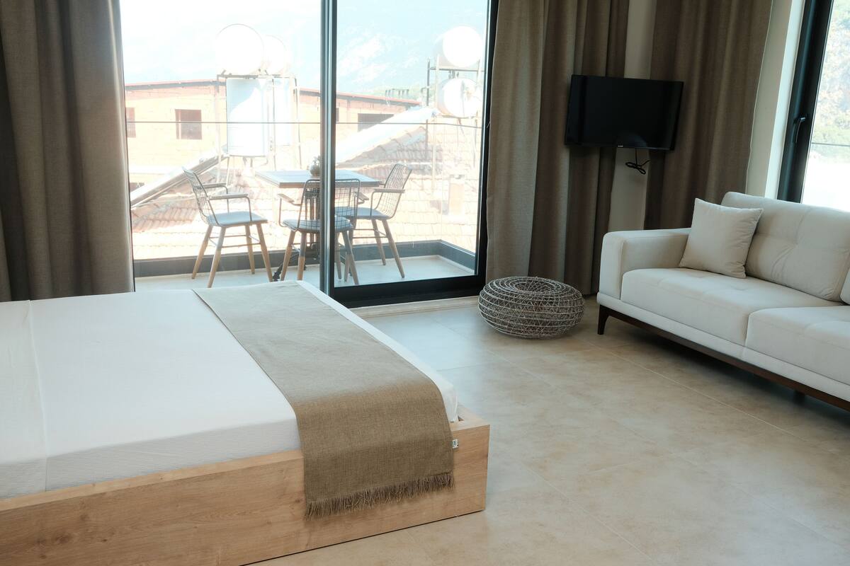 A modern hotel room with a large bed, a sofa, a TV, and a balcony with a table and chairs.