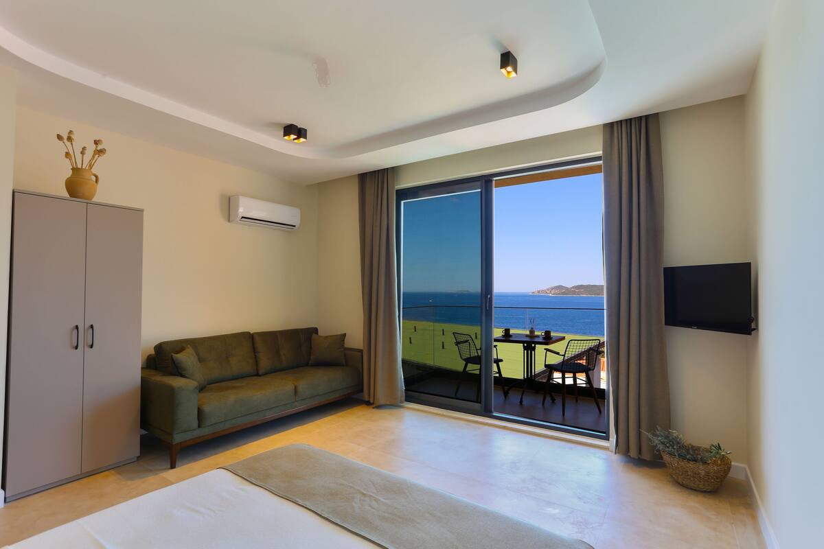 A modern living room with a sofa, a flat-screen TV, and a view of the sea through large sliding doors.