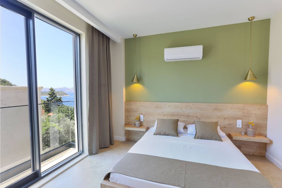 A bedroom with a large window, a bed with pillows, bedside tables, and an air conditioning unit on the wall.