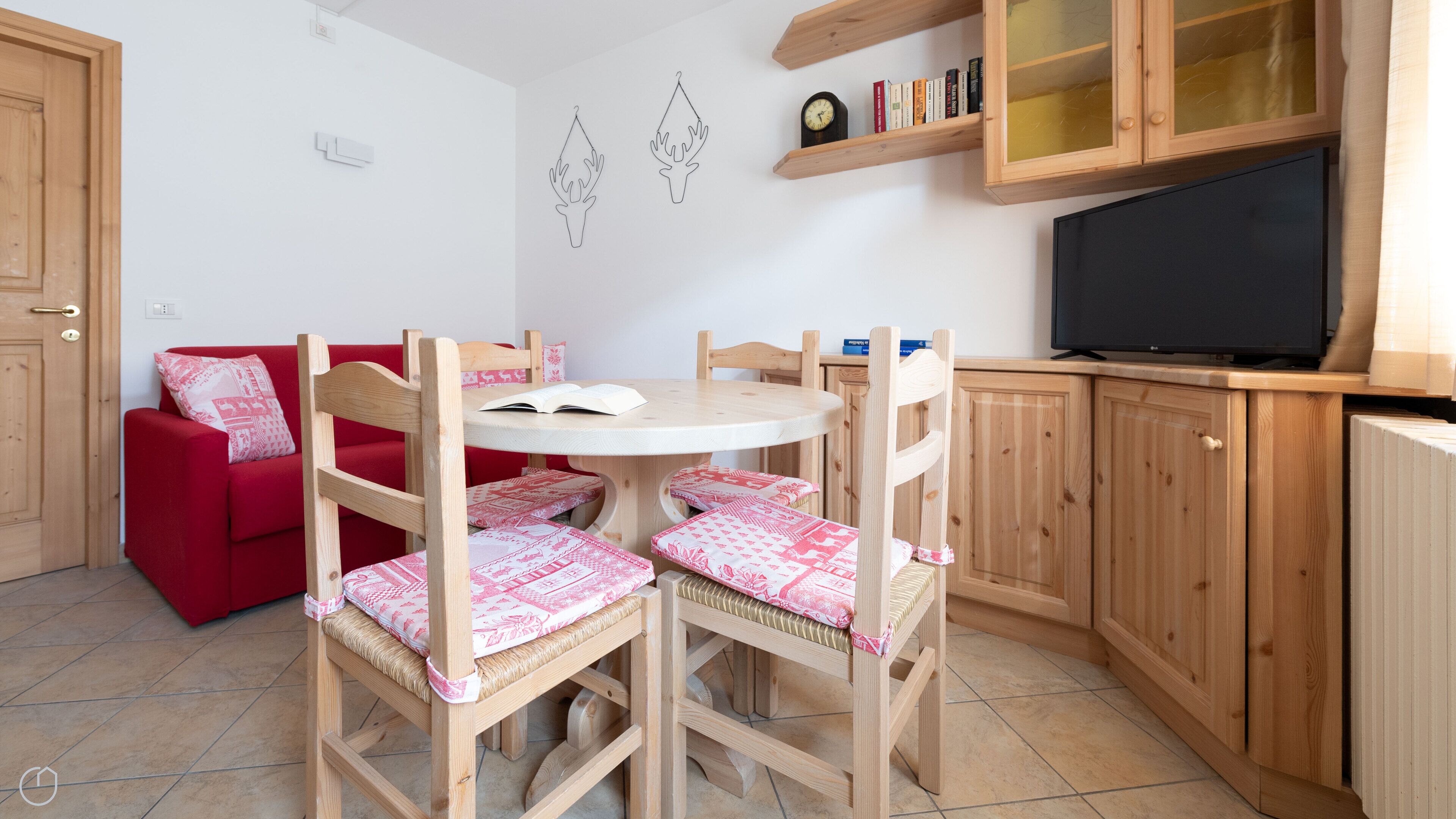 A cozy room with a red sofa, wooden chairs with patterned cushions, a round table, a television, and wooden cabinets.