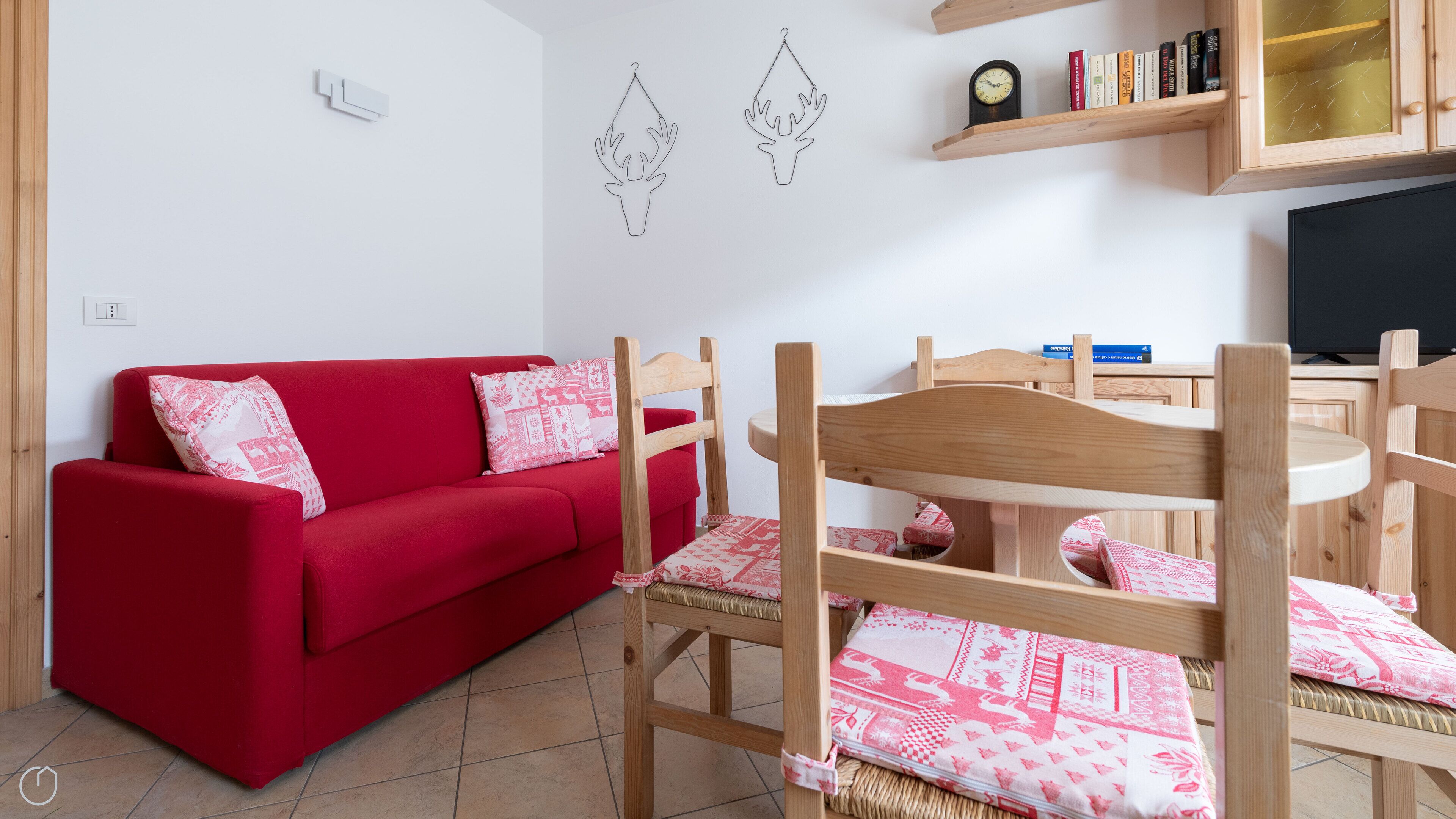 Un sofá rojo con cojines estampados, una mesa de comedor de madera con sillas, un televisor y una estantería con libros y objetos decorativos.
