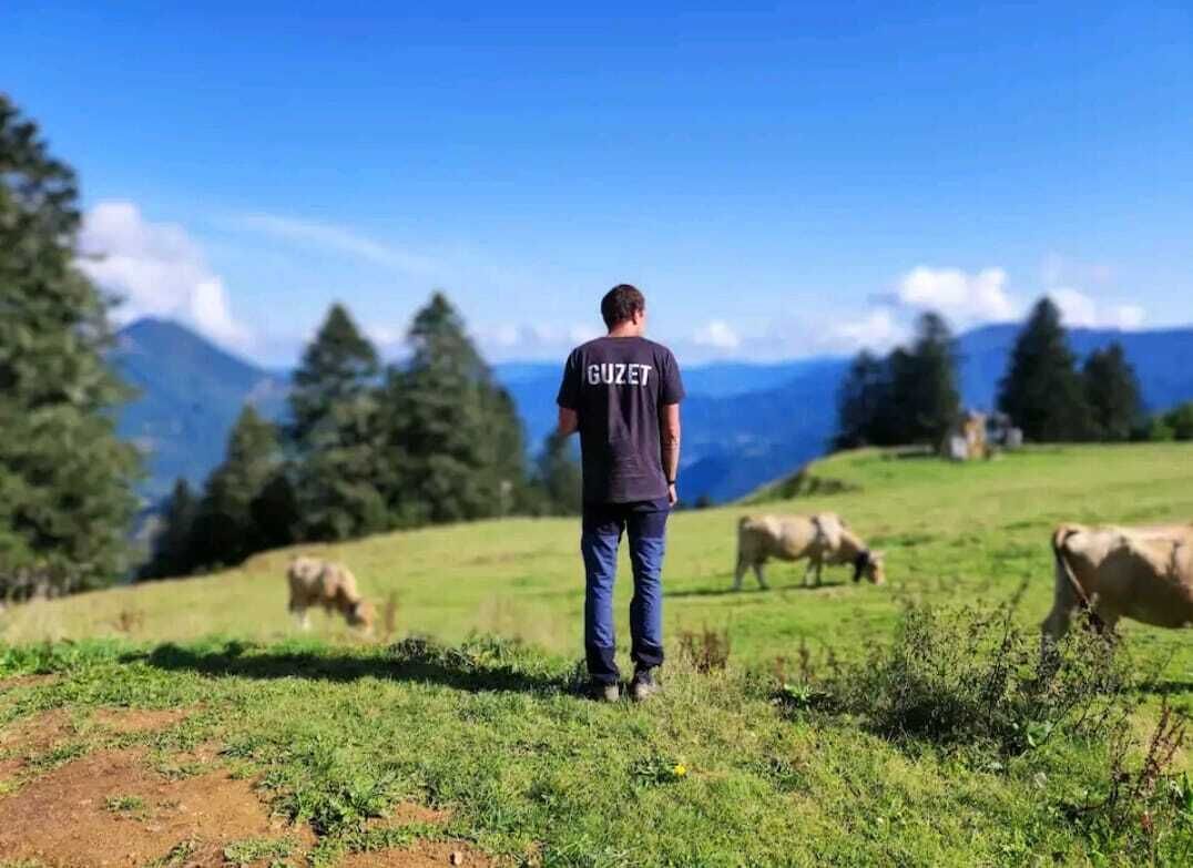 Un homme se tient debout dans un champ herbeux, avec des vaches en arrière-plan et des montagnes à l’horizon.