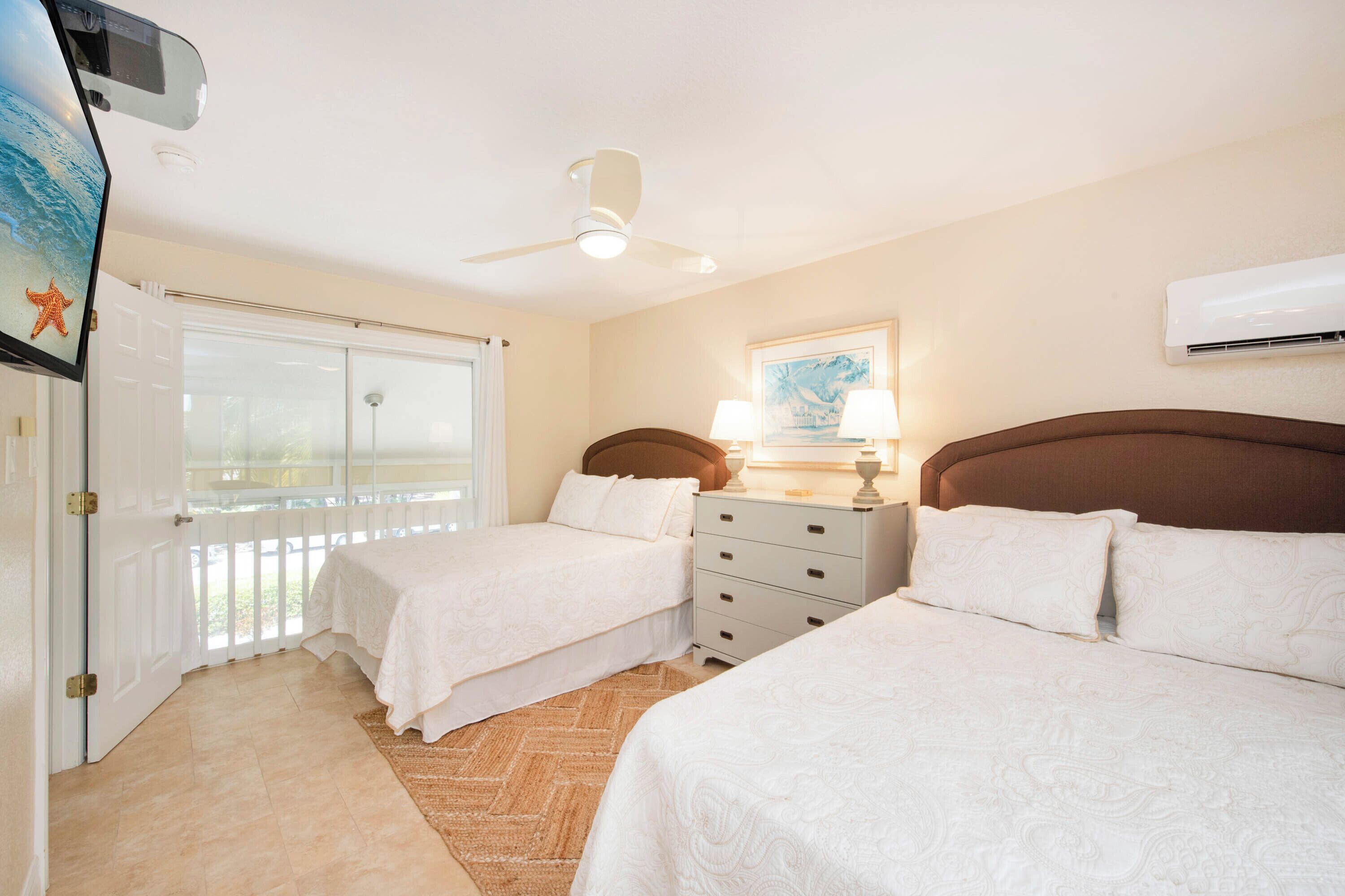 Habitación de hotel con dos camas, ventilador de techo, ventana con vistas y un cuadro en la pared.