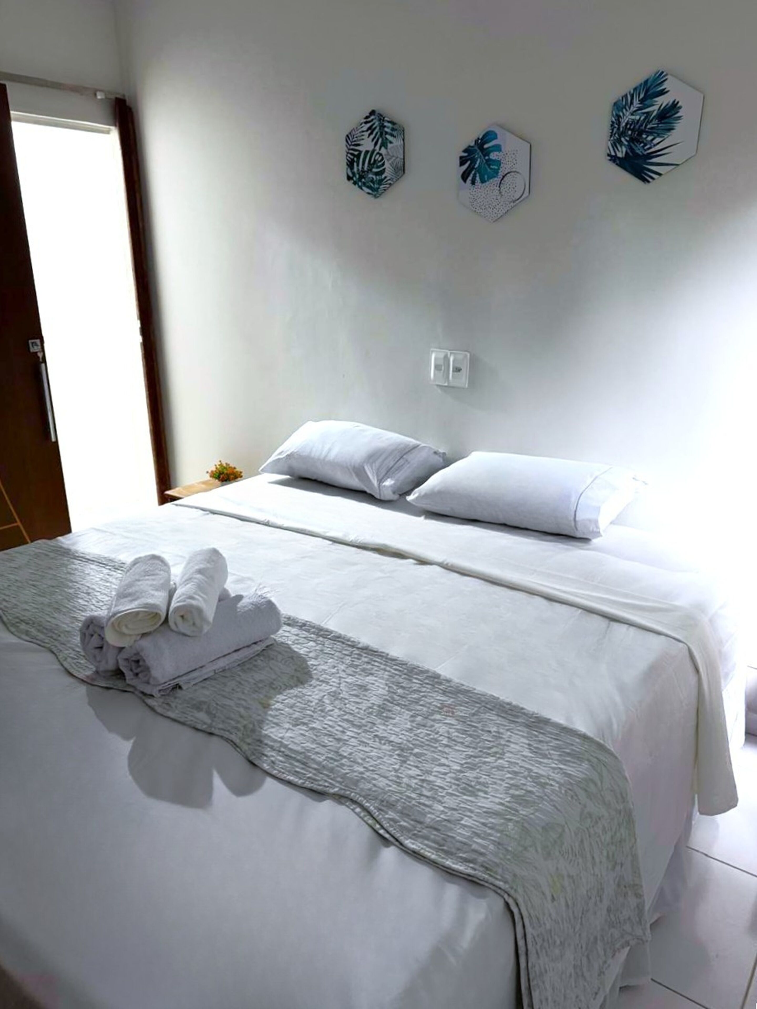 Un lit bien fait, avec du linge de lit blanc et un plaid gris, une petite table de chevet ornée d’une plante, et trois objets décoratifs accrochés au mur.
