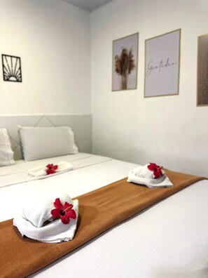 Quarto de hotel com cama arrumada, cobre marrom e duas toalhas com flores vermelhas. Três quadros pendurados na parede.