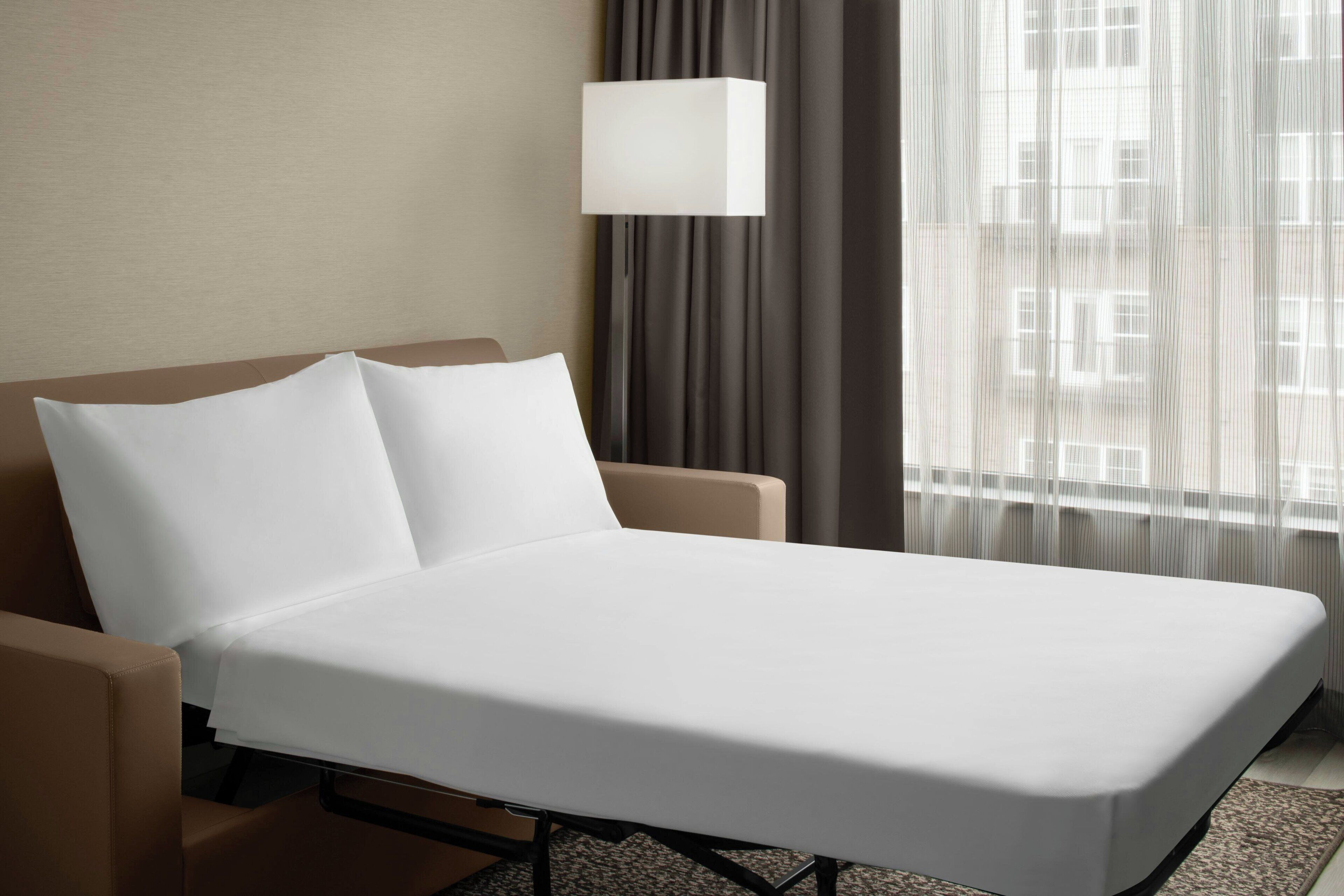 A hotel room with a bed, a chair, a lamp, and a window with curtains.