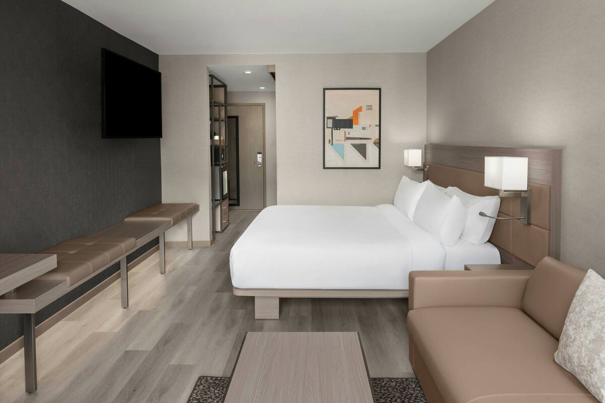 A modern hotel room with a bed, a sofa, a bench, a flat-screen TV, and a framed artwork on the wall.