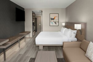 A modern hotel room with a bed, a sofa, a bench, a flat-screen TV, and a framed artwork on the wall.