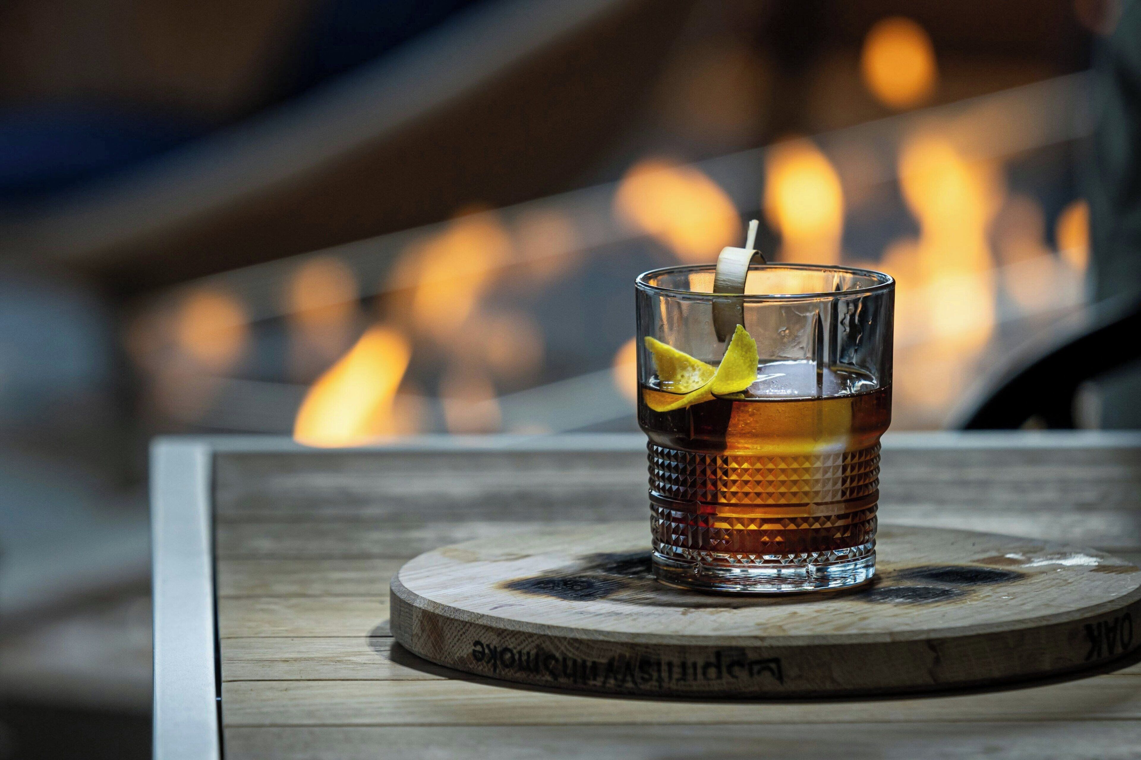 A glass of amber-colored beverage with a lemon slice garnish on a wooden coaster with engraved text, placed on a table with a blurred background of warm lighting.
