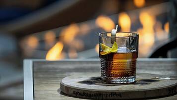 A glass of amber-colored beverage with a lemon slice garnish on a wooden coaster with engraved text, placed on a table with a blurred background of warm lighting.