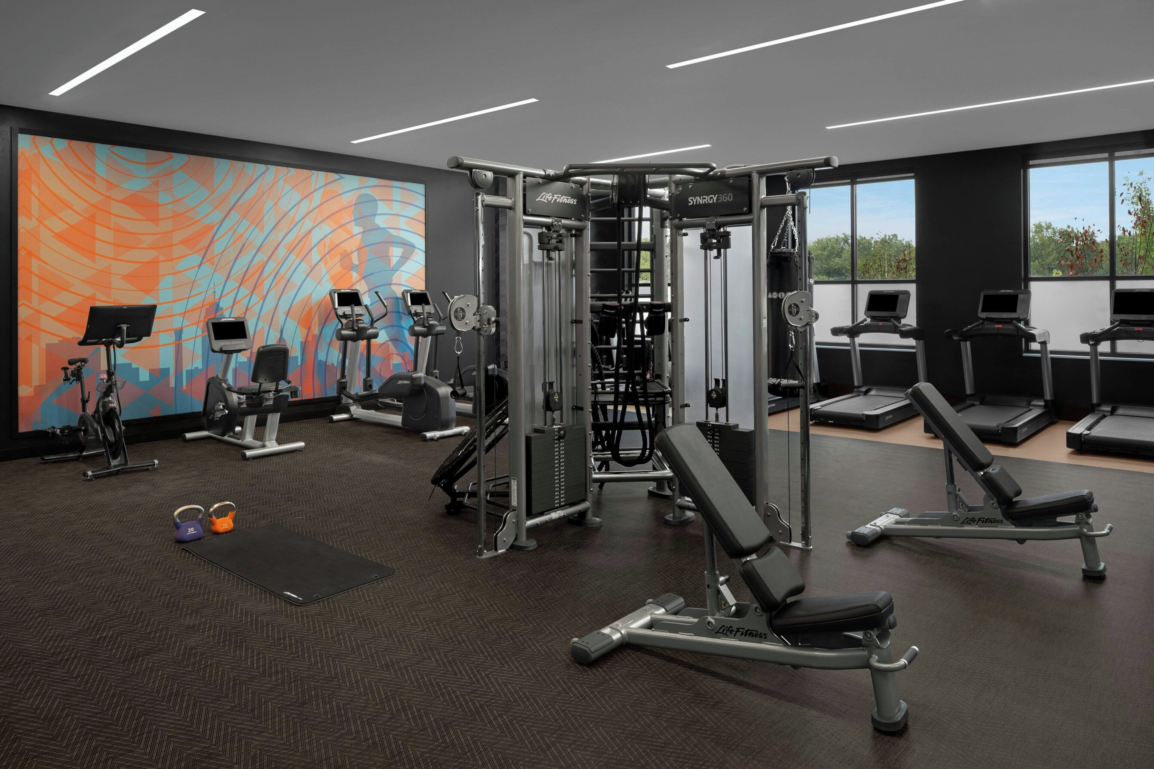 Een moderne sportschool met diverse fitnessapparaten, grote ramen en een kleurrijke muurschildering.
