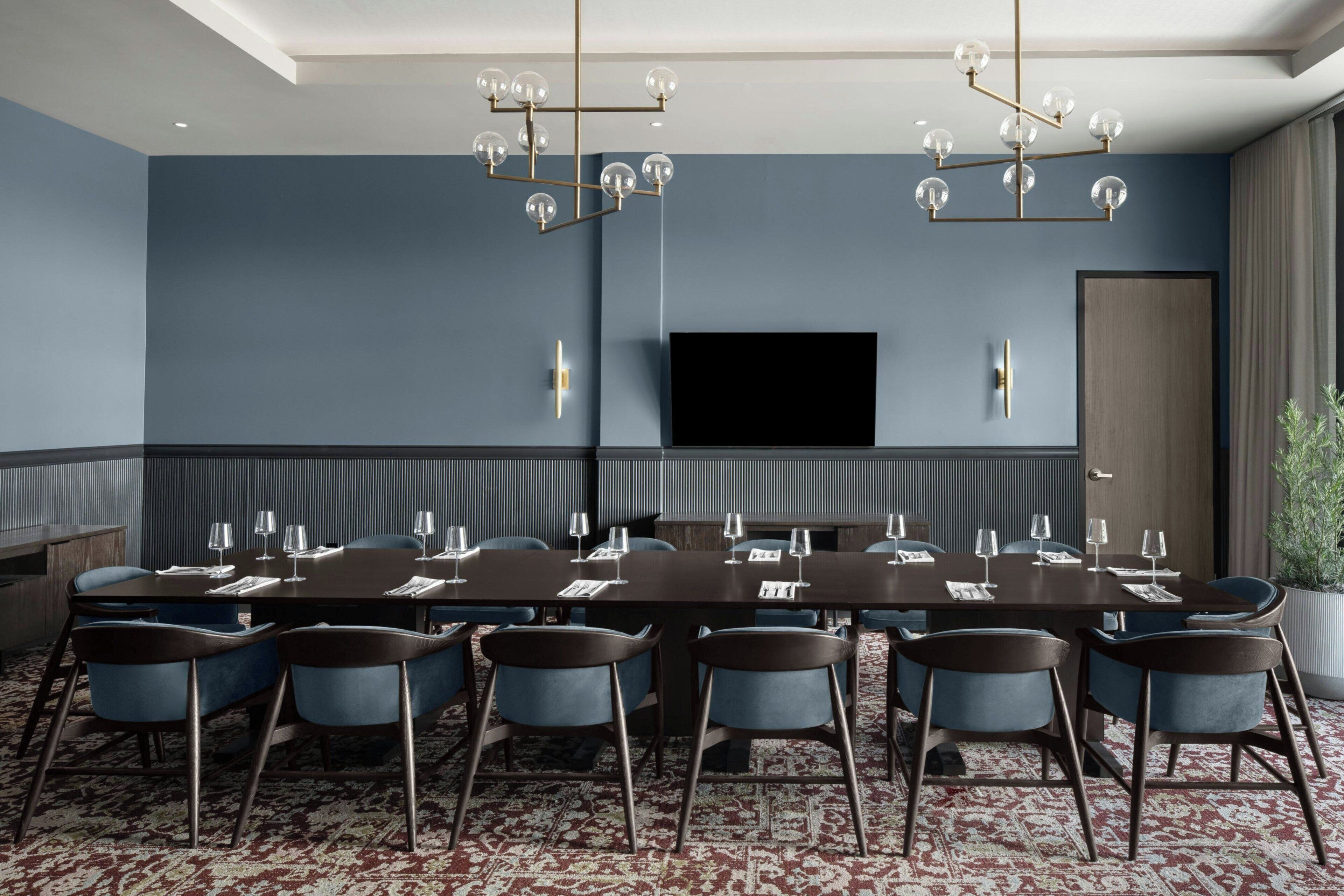 A conference room with a long table, blue chairs, a chandelier, a flat-screen TV, and a potted plant.