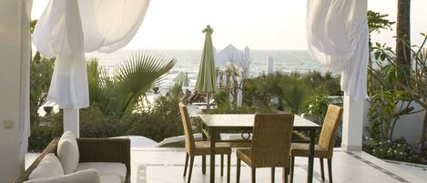 Un área de comedor al aire libre con techo, mobiliario de mimbre, una mesa y sillas, con vistas al océano y palmeras.