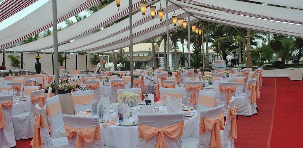 A banquet hall with round tables covered in white tablecloths and peach-colored sashes, arranged under a white canopy with hanging lights.