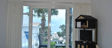 A hotel room with a bed, a TV, and a balcony with a view of palm trees and a building.
