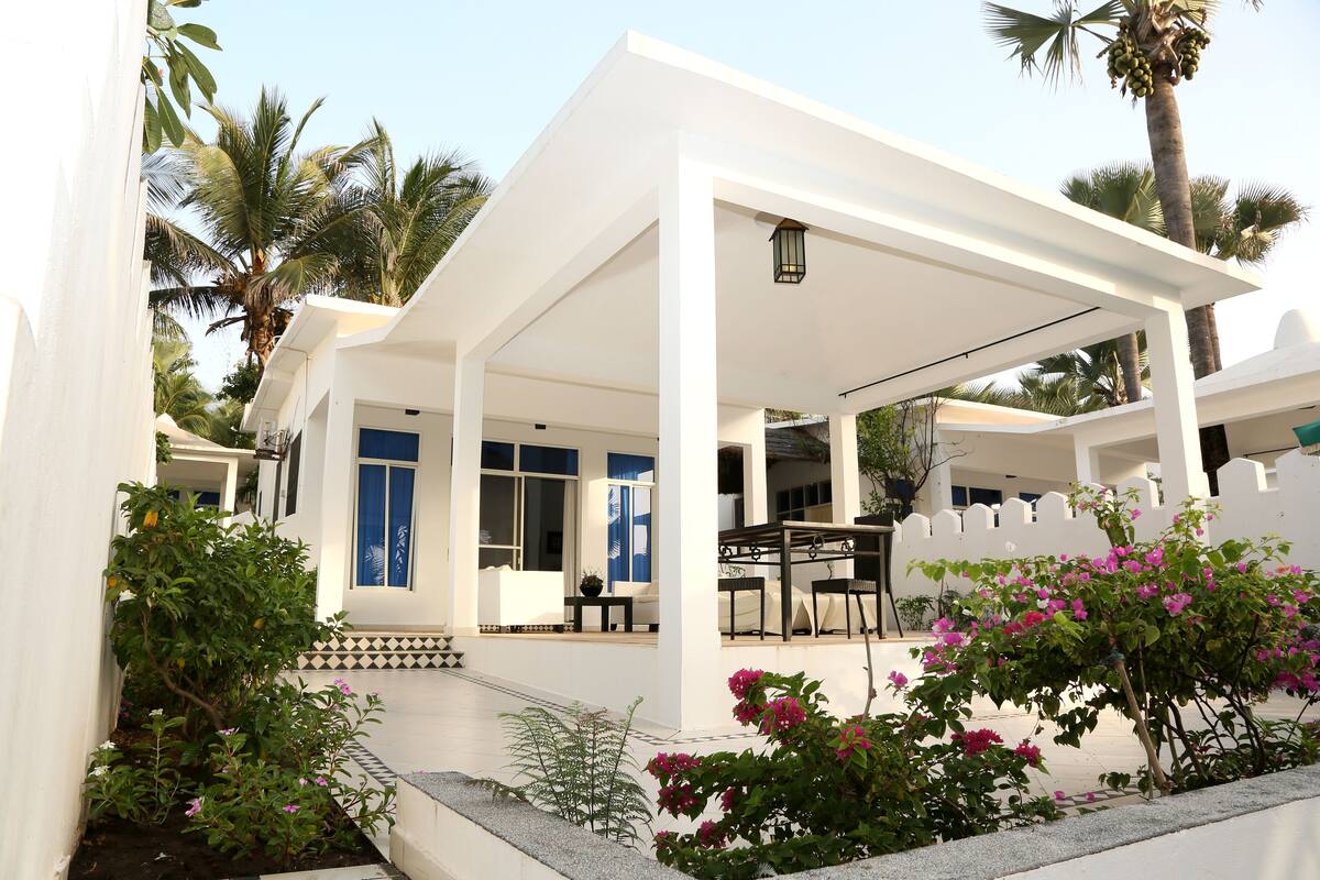 A white building with a covered patio area, palm trees, and flowering plants.