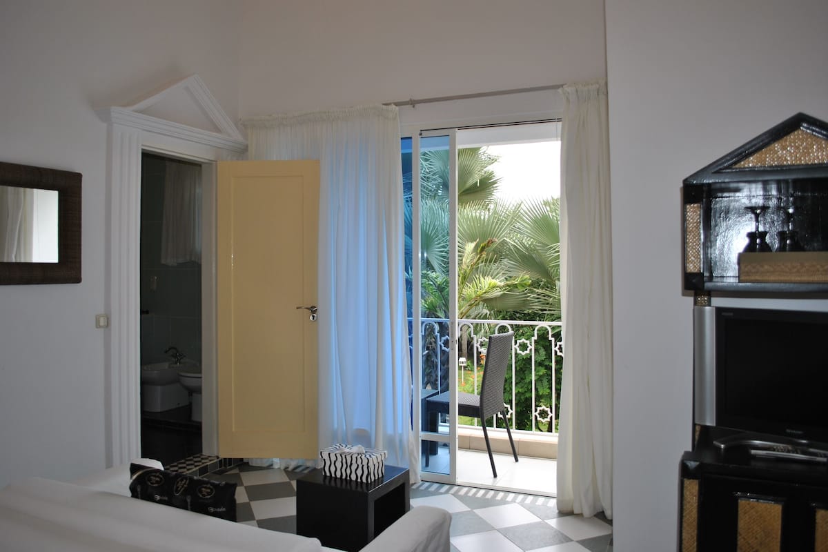 A hotel room with a checkered floor, a balcony with a view of palm trees, a small table with a decorative item, and a television.