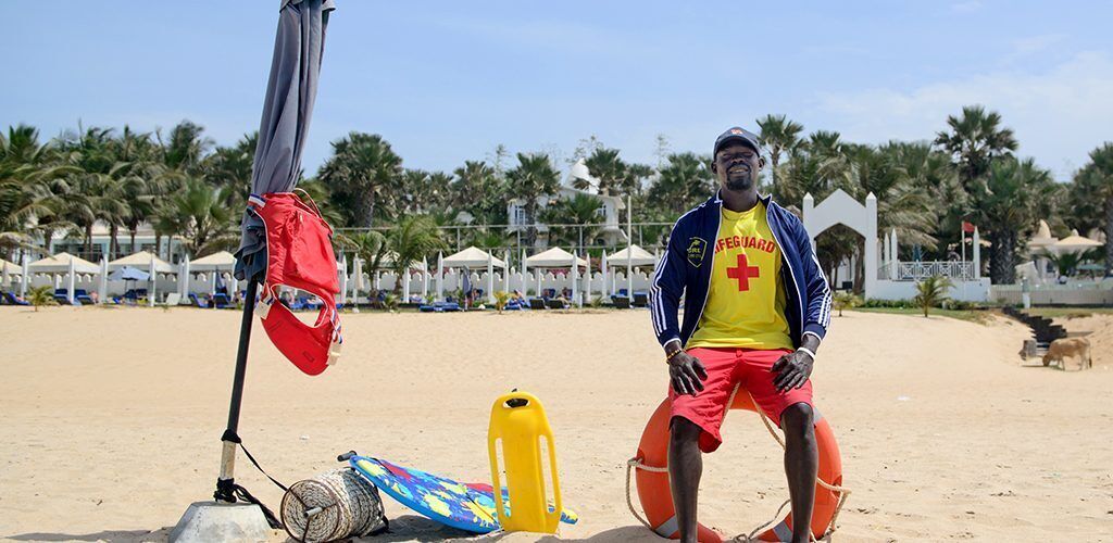 A lifeguard on a sandy beach with beach umbrellas and palm trees in the background.