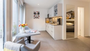 Apartament modern i compacte amb cuina americana, zona de menjador amb taula i cadires, finestra gran i un llit a l'habitació veïna.