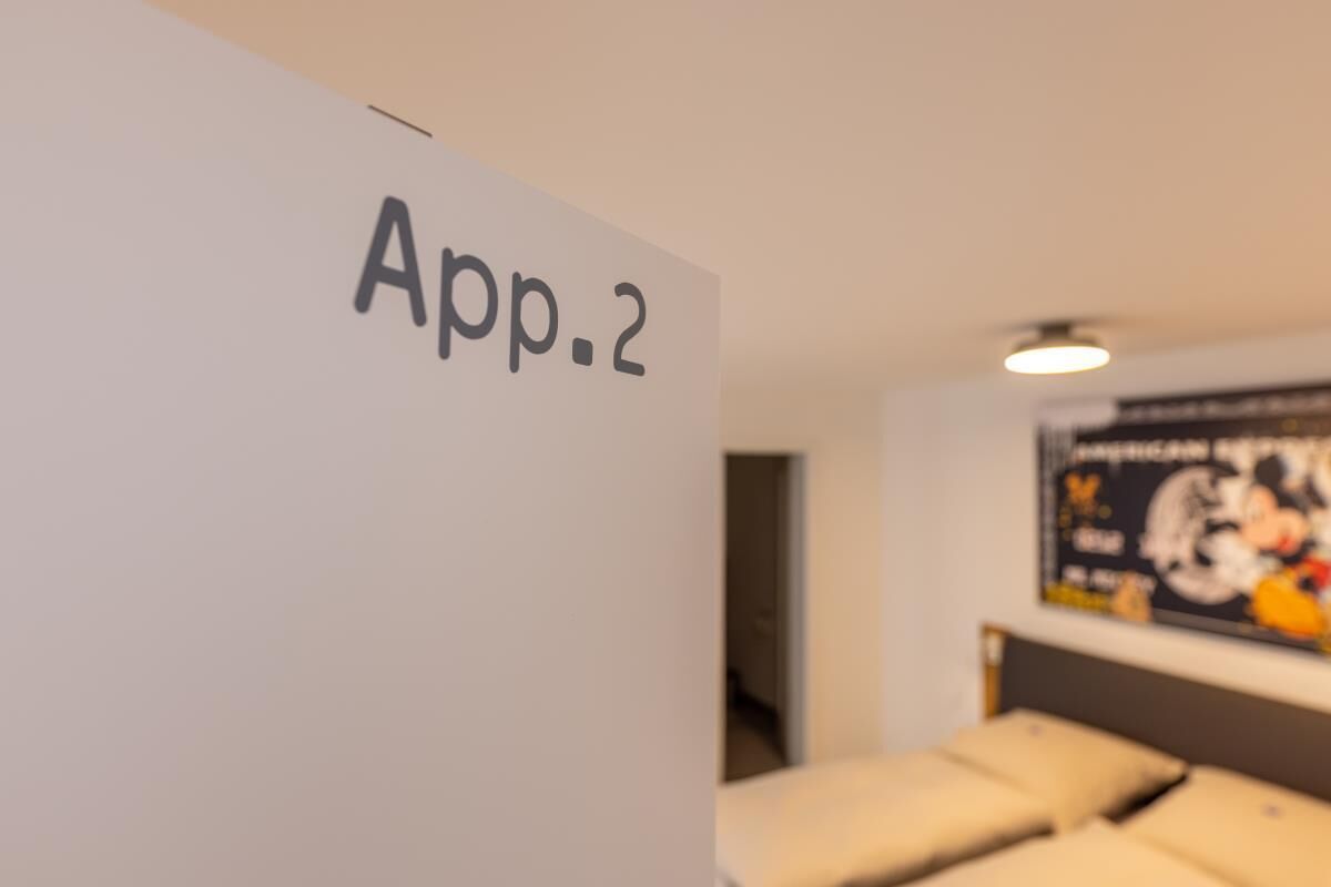 Eine Wand mit der Aufschrift 'App.2', ein Bett mit Kissen und ein Poster an der Wand.